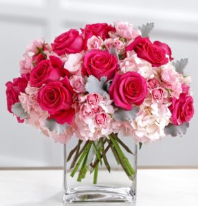 A delicate and sophisticated bouquet of sweet feminine flowers. Bright fuchsia roses are brilliantly eye-catching seated amongst pale pink spray roses and clouds of pink blush hydrangea accented with floral greens. Elegantly situated in a clear glass vase with a modern shape,