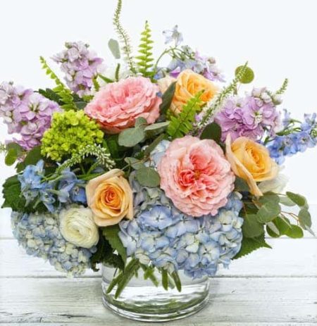 Inspire with a bouquet of the English morning countryside. Dreamy pastels of fragrant lavender stock, delicate blue delphiniums, pillowy Italian ranunculus dance with willows of of veronica in a bed of English blush pink garden roses, sun-bleached hydrangea and silver eucalyptus. Hand designed in a farm to table gathering vase its perfectly presented as a table centerpiece, credenza or desktop display of timeless floral perfection.
