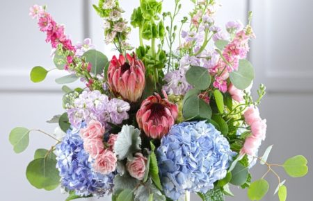 Pastel garden favorites including southern blue hydrangea, fragrant stock, wild pink tea roses, bells of Ireland, larkspur and snapdragons, pink mink protea, and lush floral foliage presented in a chic glass vase.
