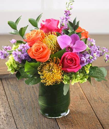 The Mosaic features fragrant purple stock, hot pink and sunset orange mountain roses, exotic pincushion protea and orchids for a vibrant, bright and cheery presentation artfully arranged in a leaf-wrapped glass vase.