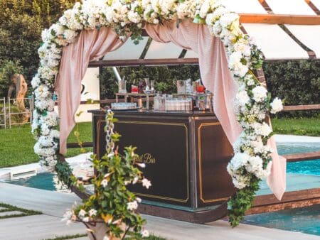 A beautifully decorated outdoor bar setup featuring abundant white floral arrangements and soft drapery, perfect for weddings or parties